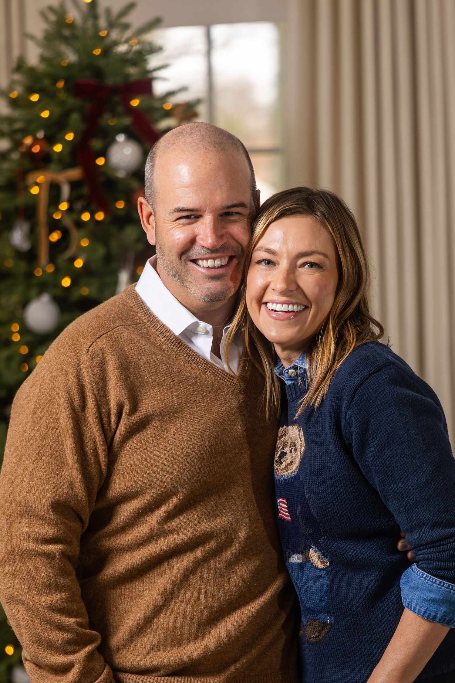 A couple poses together in front of a decorated Christmas tree wearing brown and navy sweaters.