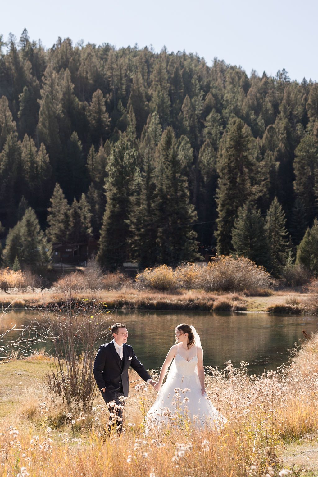 A couple stands in a golden meadow beside a serene lake surrounded by evergreen forest on a sunny autumn day.