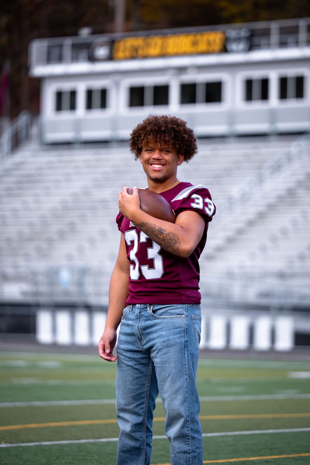 Child in maroon sports jersey and jeans plays on football field with stadium seating visible.