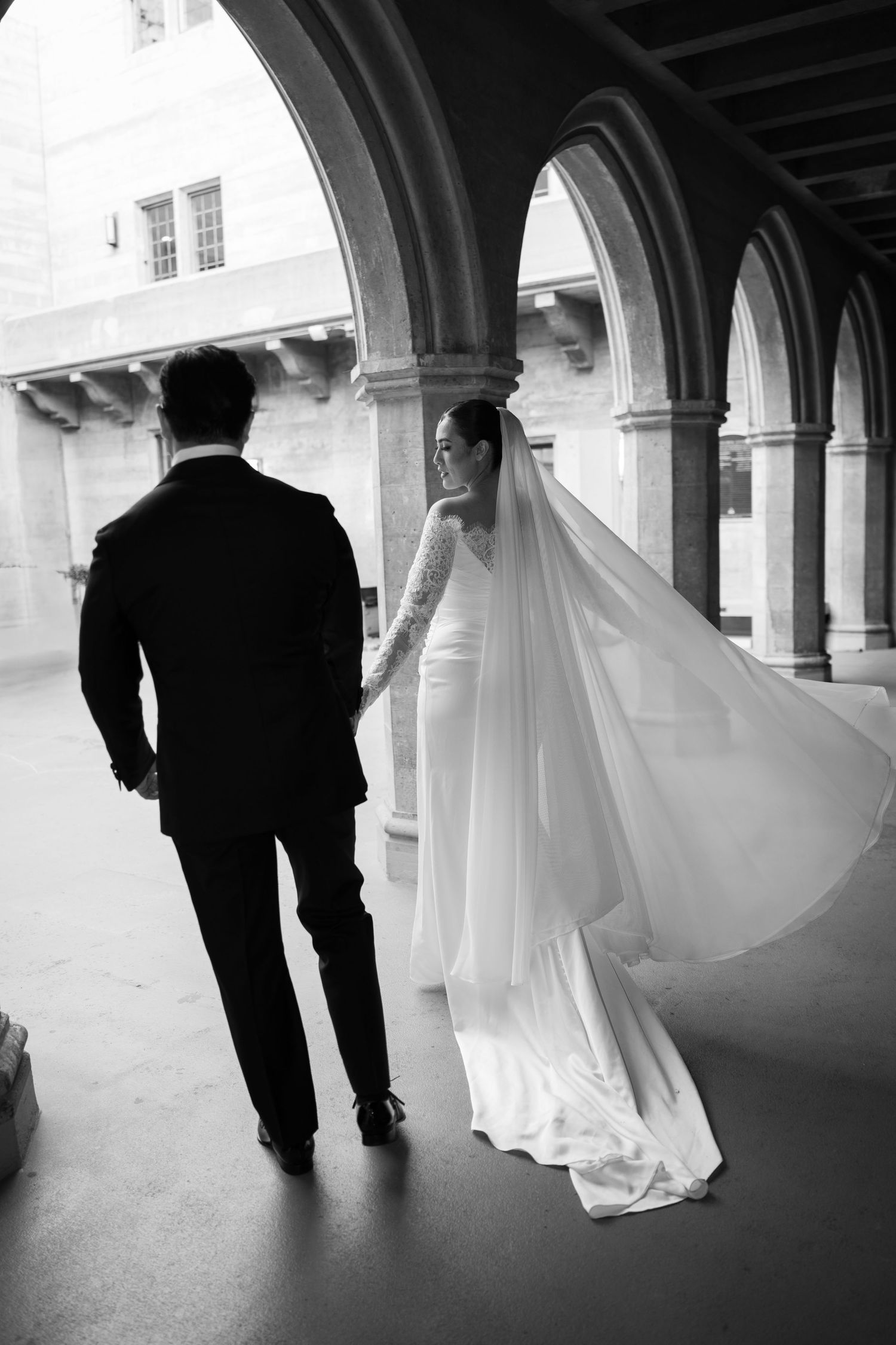 A black and white wedding photo series showing silhouettes of a couple walking under arched columns with flowing veil.