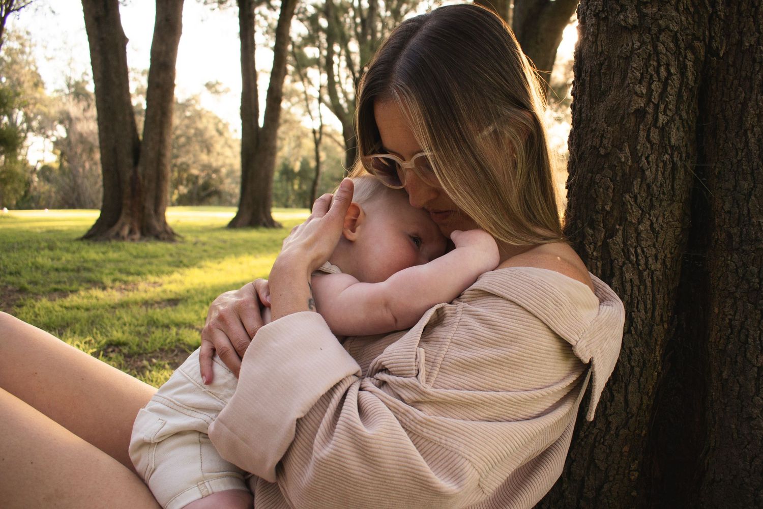 mother holds her baby close as she leans against a tree during a motherhood breastfeeding milestone photoshoot Maitland NSW