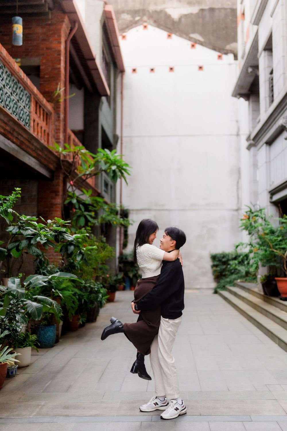 Two figures share an intimate moment in a narrow alleyway lined with potted plants and traditional architecture.