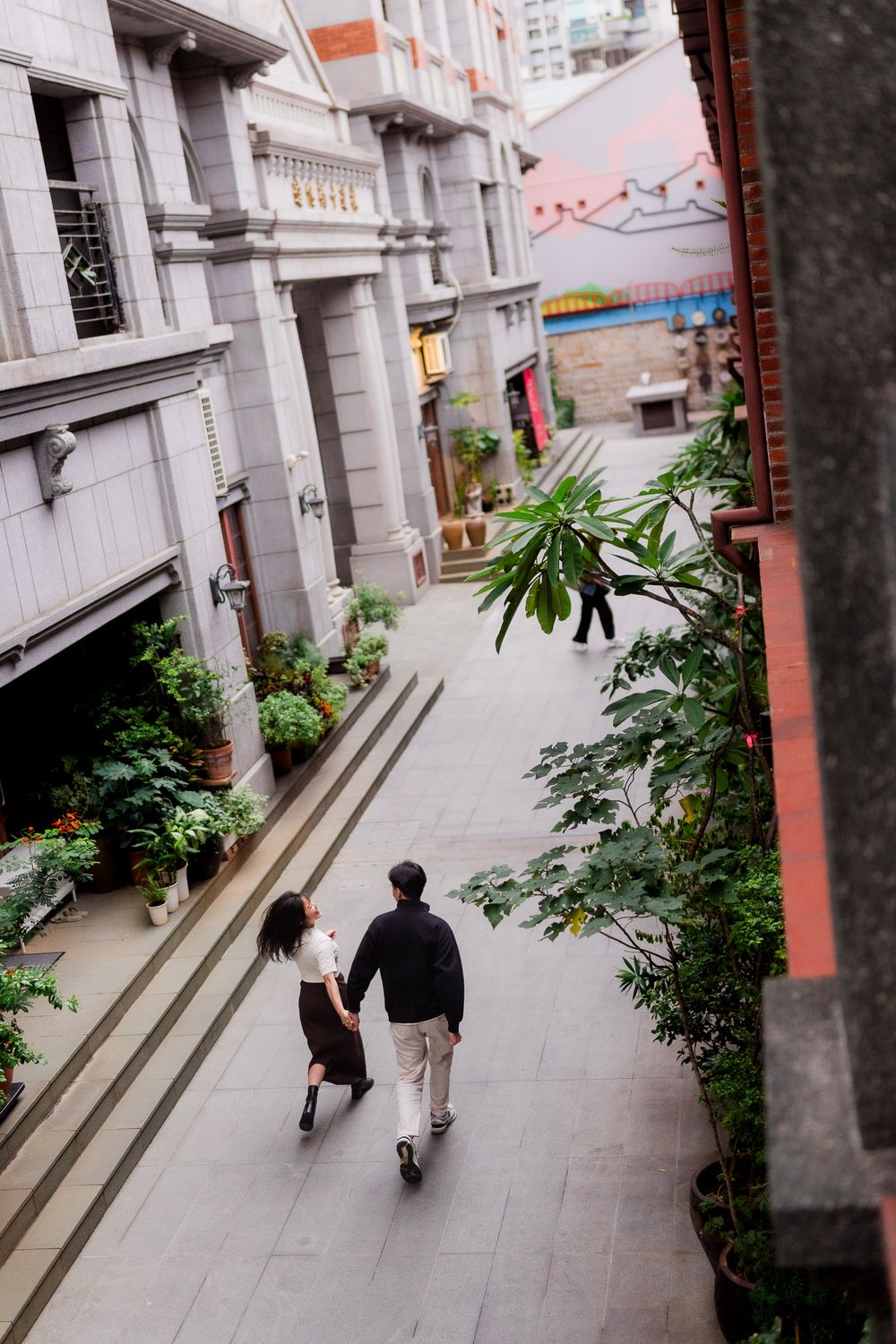 People walking along a modern urban corridor with white walls and plants lining the pathway.