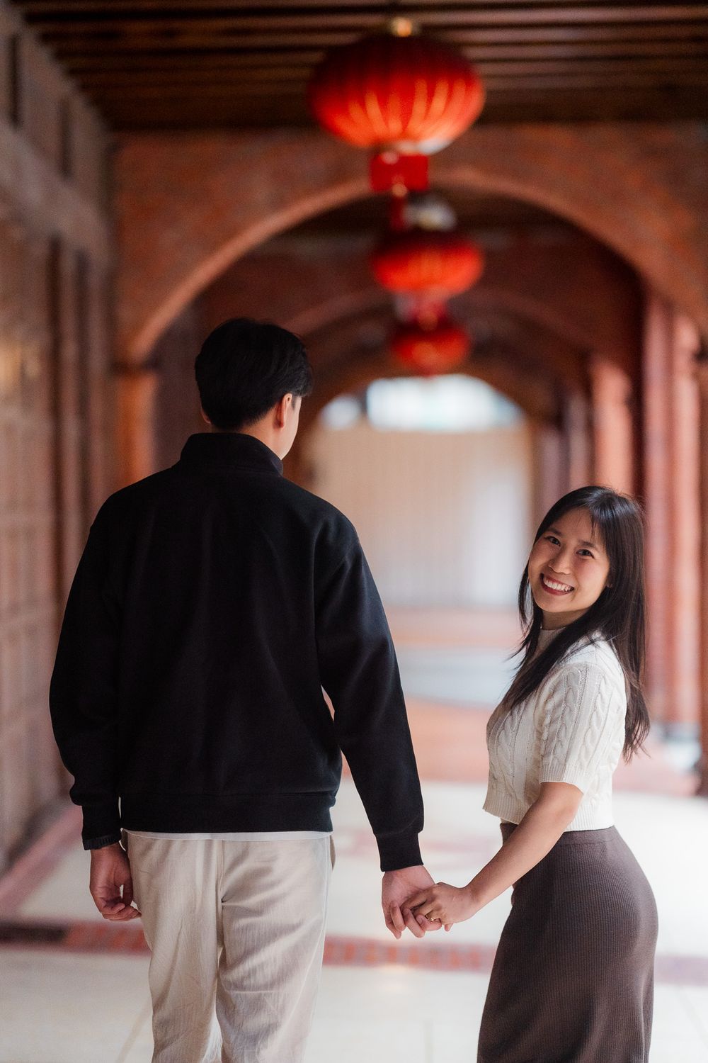 Couple holding hands in hallway with traditional red Chinese lanterns overhead creating romantic ambiance.