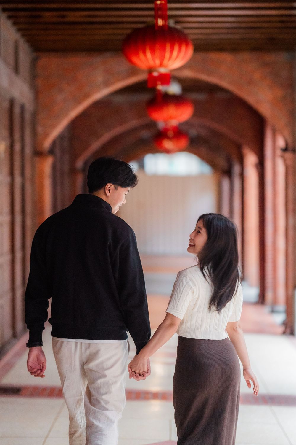 A couple walks hand in hand through a corridor with traditional red Chinese lanterns overhead.
