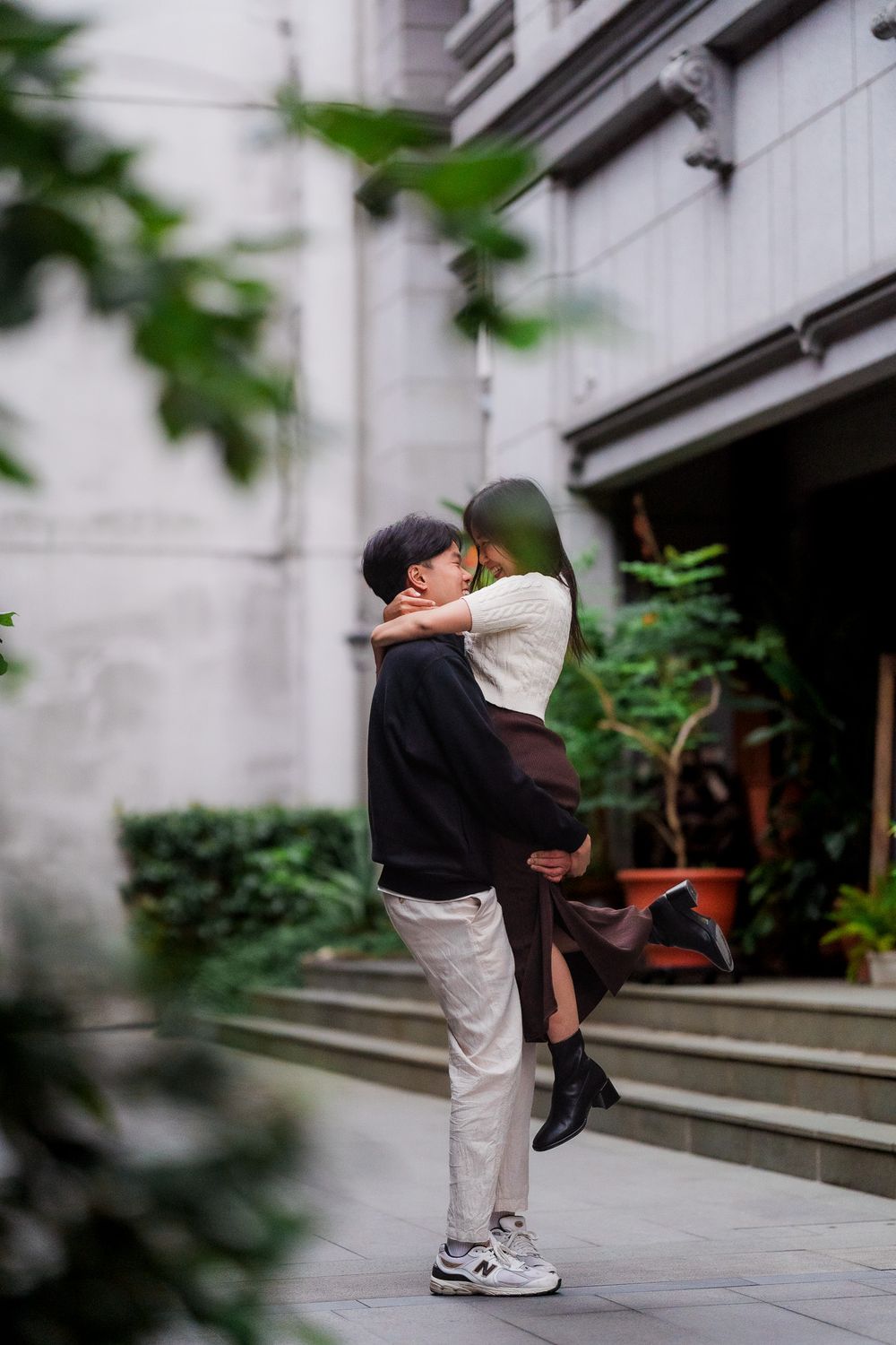 A couple embraces on stone steps outside a building, surrounded by green foliage in a romantic moment.