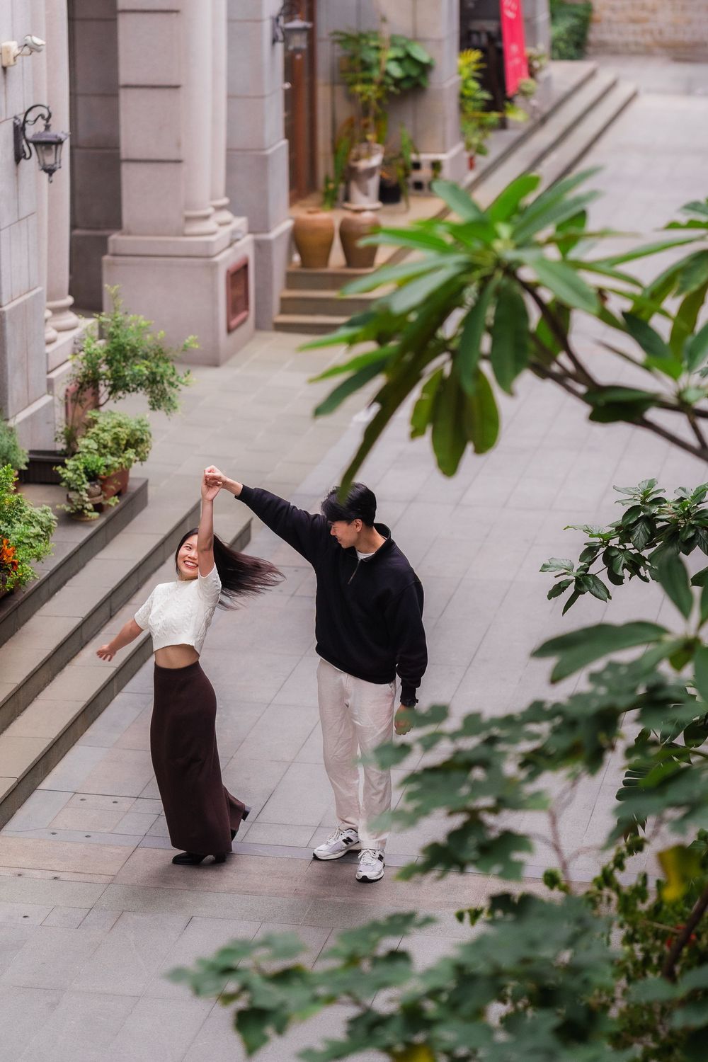Two people practice martial arts kicks in a sequence along a building exterior with potted plants.