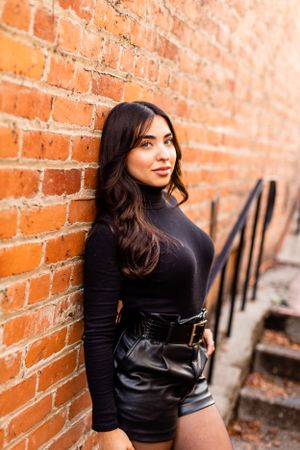 Portrait against orange brick wall featuring black turtleneck and leather shorts with belt.