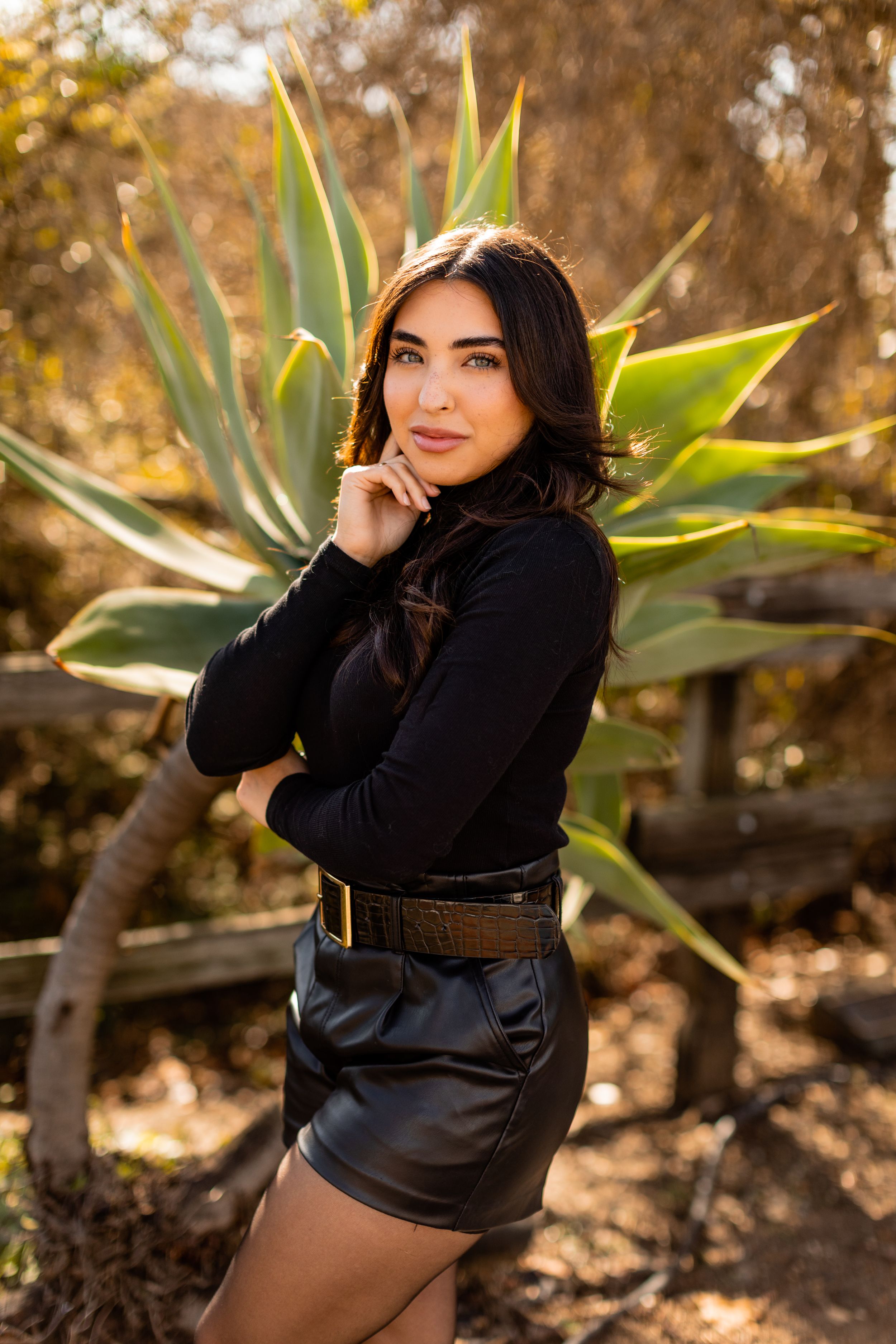 A fashion portrait against agaves features a black turtleneck and leather shorts in warm golden lighting.