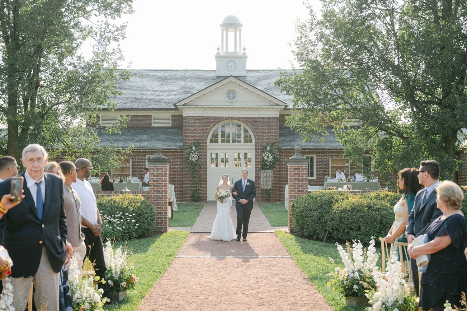Wedding party walks through a garden path towards a traditional brick church building with white columns and steeple. - Elawa Farm