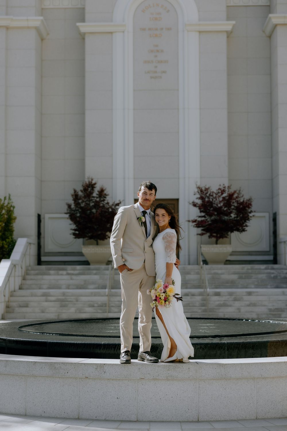 A couple in wedding attire poses together in front of a white temple building with a fountain in the foreground.