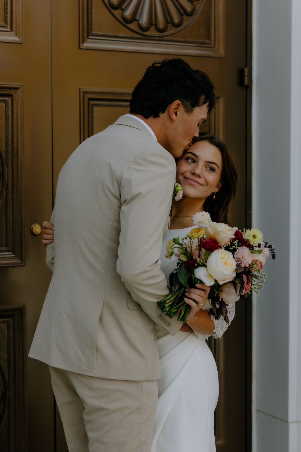 A romantic wedding couple shares tender moments at a doorway with a beautiful floral bouquet.