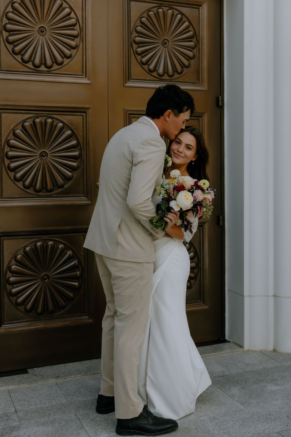 A couple in wedding attire poses for romantic photos in front of ornate wooden doors with sunburst patterns.