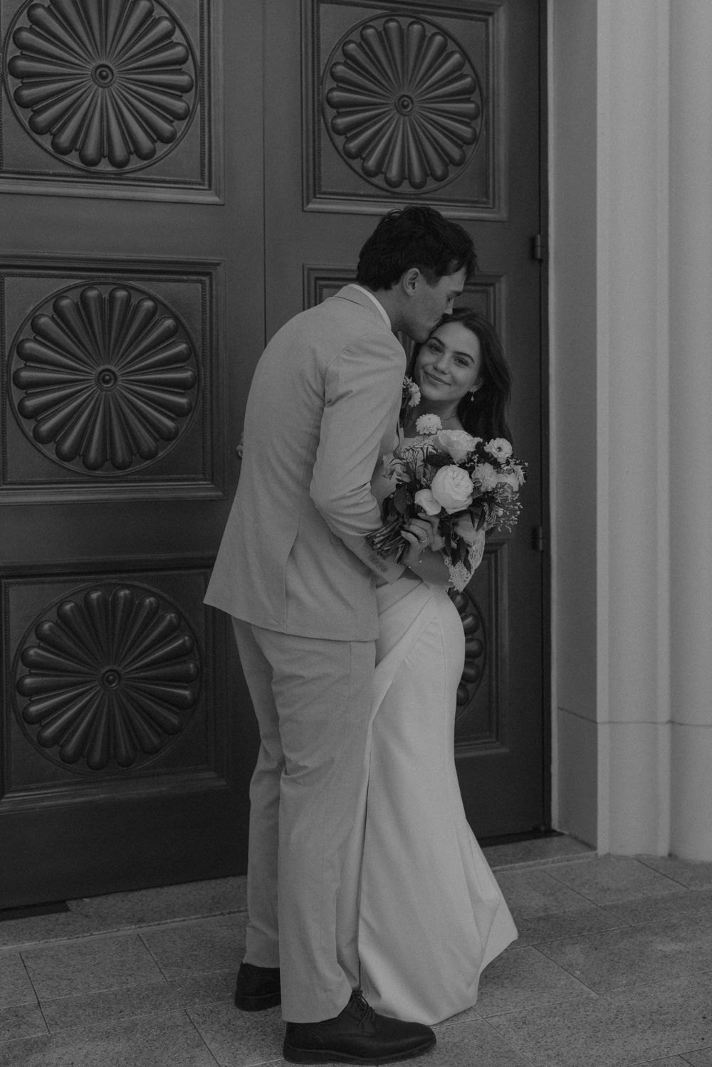 A couple in wedding attire shares an intimate moment in front of ornate wooden doors with sunburst patterns.