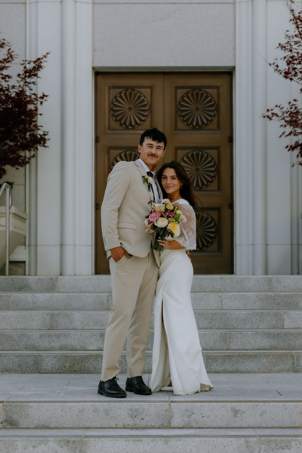 Wedding couple stands on stone steps in front of decorative brown doors with sunburst patterns.