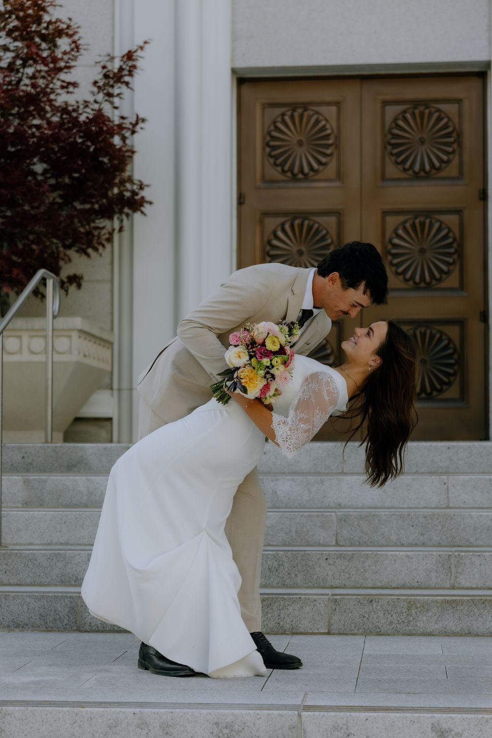 A couple in wedding attire performs a dramatic dip pose on stone steps in front of ornate wooden doors.