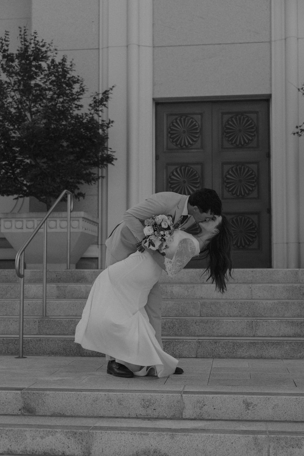 A romantic dip kiss on stone steps in front of ornate wooden doors, captured in black and white.
