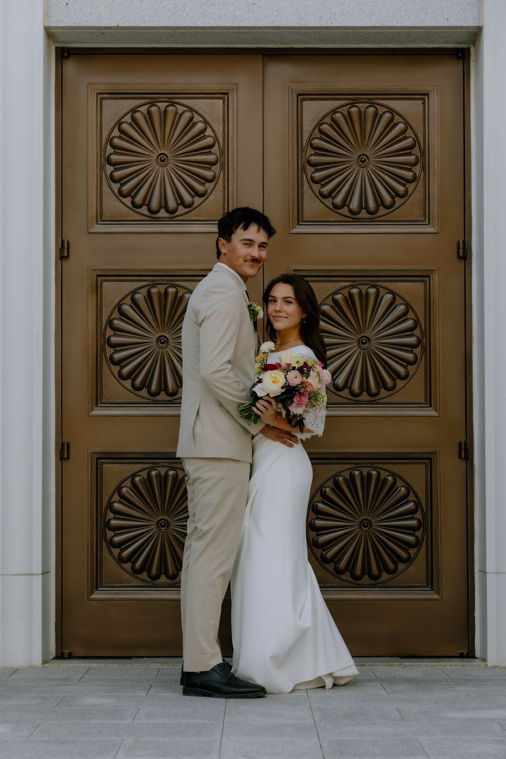 A couple in wedding attire poses against ornate wooden doors with fan-shaped designs at a historic building entrance.