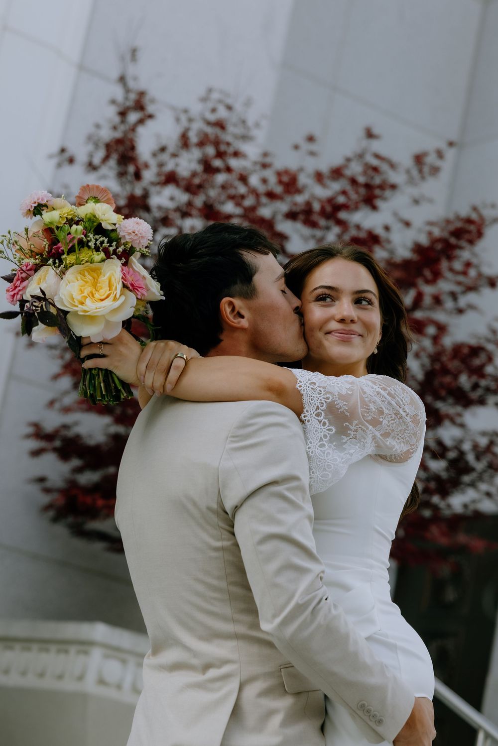 A romantic wedding couple embraces outdoors in front of red foliage while holding a floral bouquet.