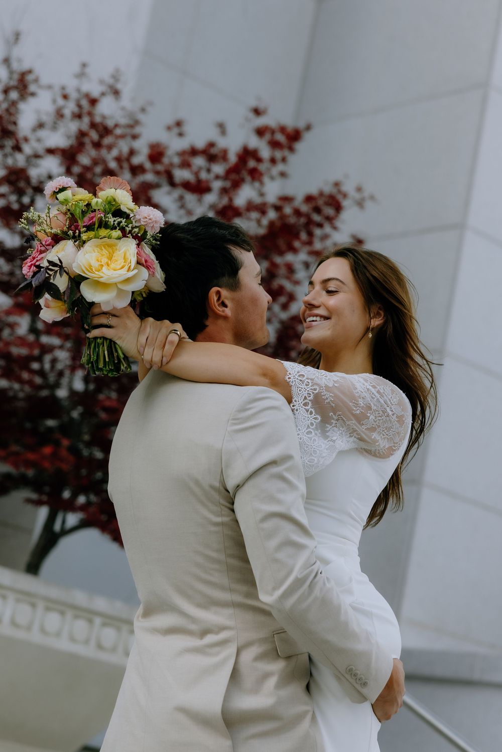 A romantic wedding couple embraces and spins joyfully in front of a white building with autumn foliage.