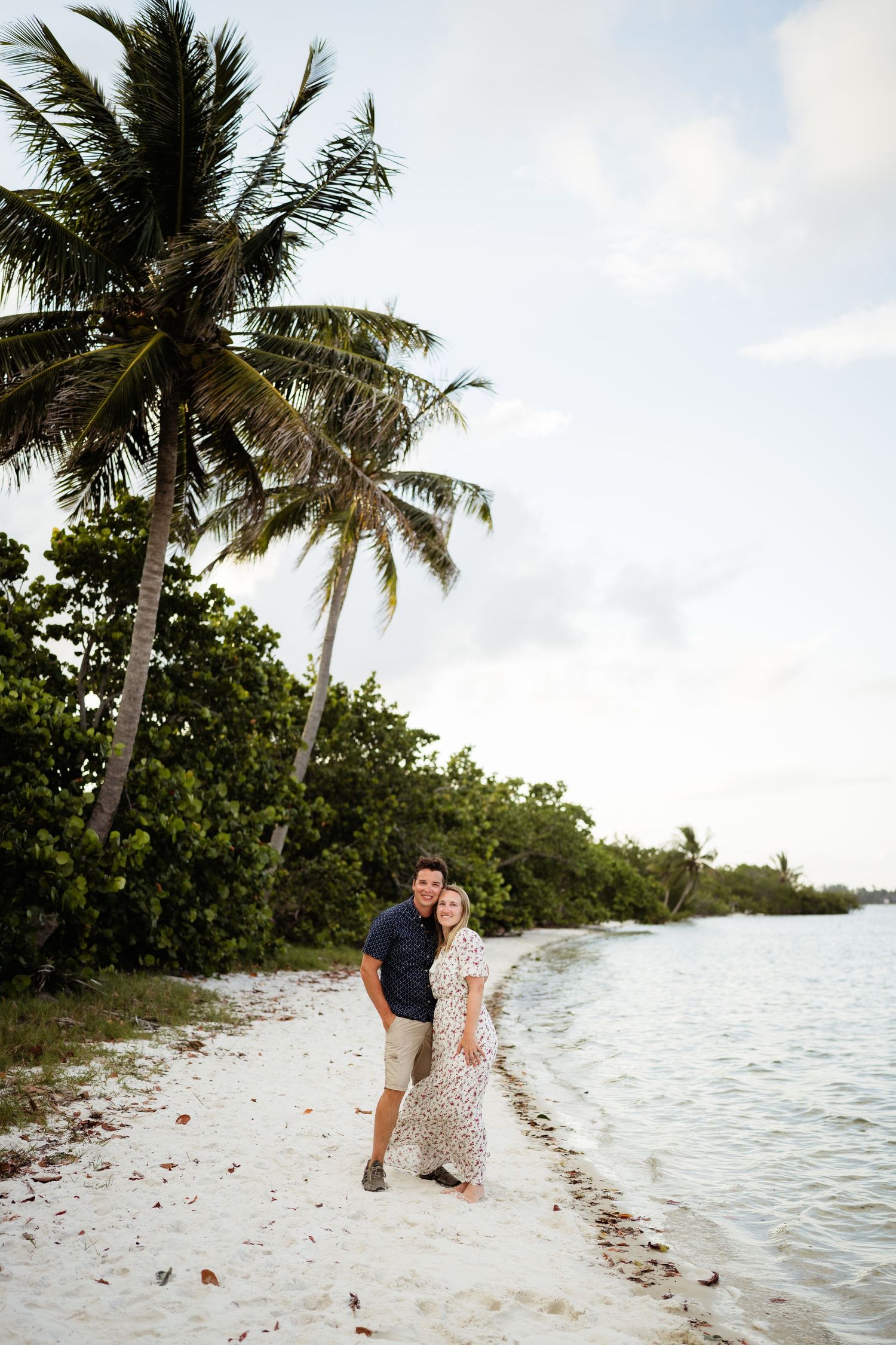 A couple embraces on a sandy beach with grass in the background on a sunny day.