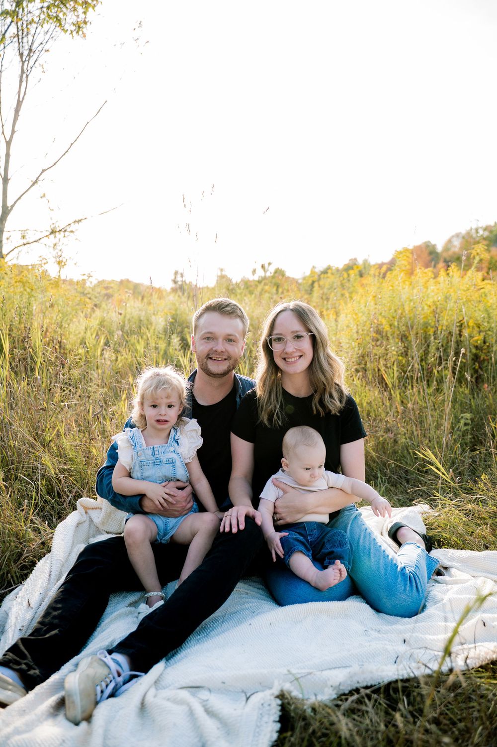 A family sits together on a white blanket in a grassy field during golden hour.
