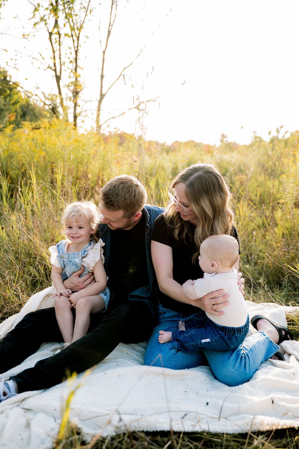 Family relaxing together on a white blanket in a sunlit meadow during golden hour.