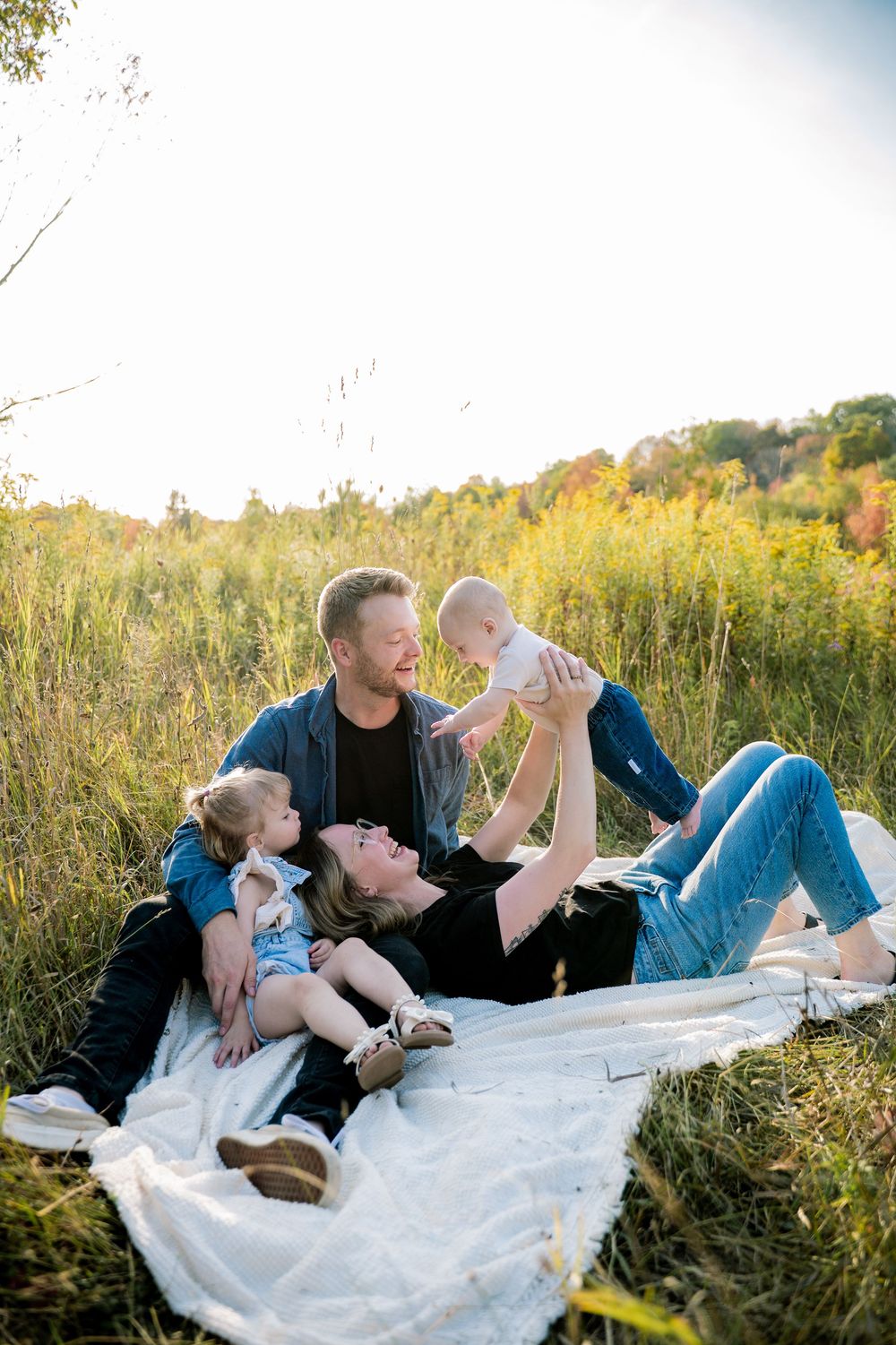 A couple relaxes on a white blanket in a sunny meadow during golden hour, surrounded by tall grass and autumn foliage.