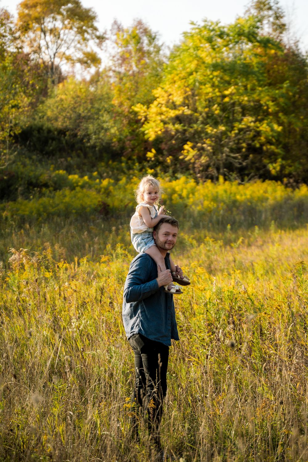 Two figures stand in a sunlit autumn meadow surrounded by tall grass and golden trees in the background.