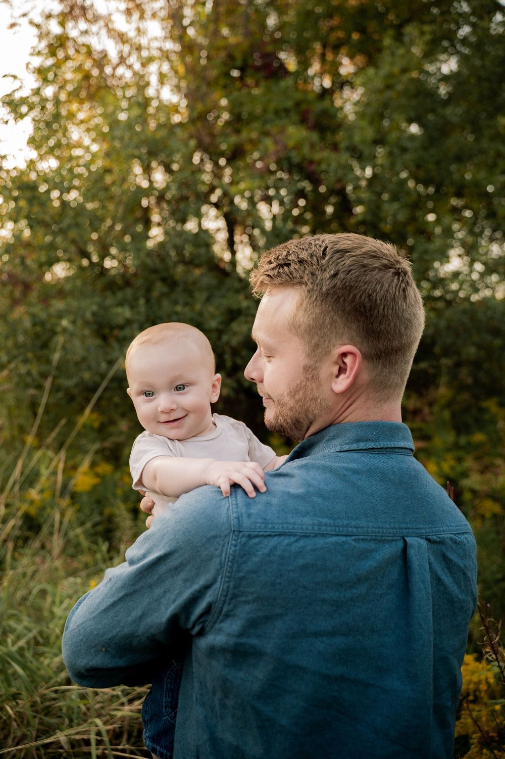 A tender outdoor portrait showing an adult in a blue denim shirt holding a baby against a natural green background.