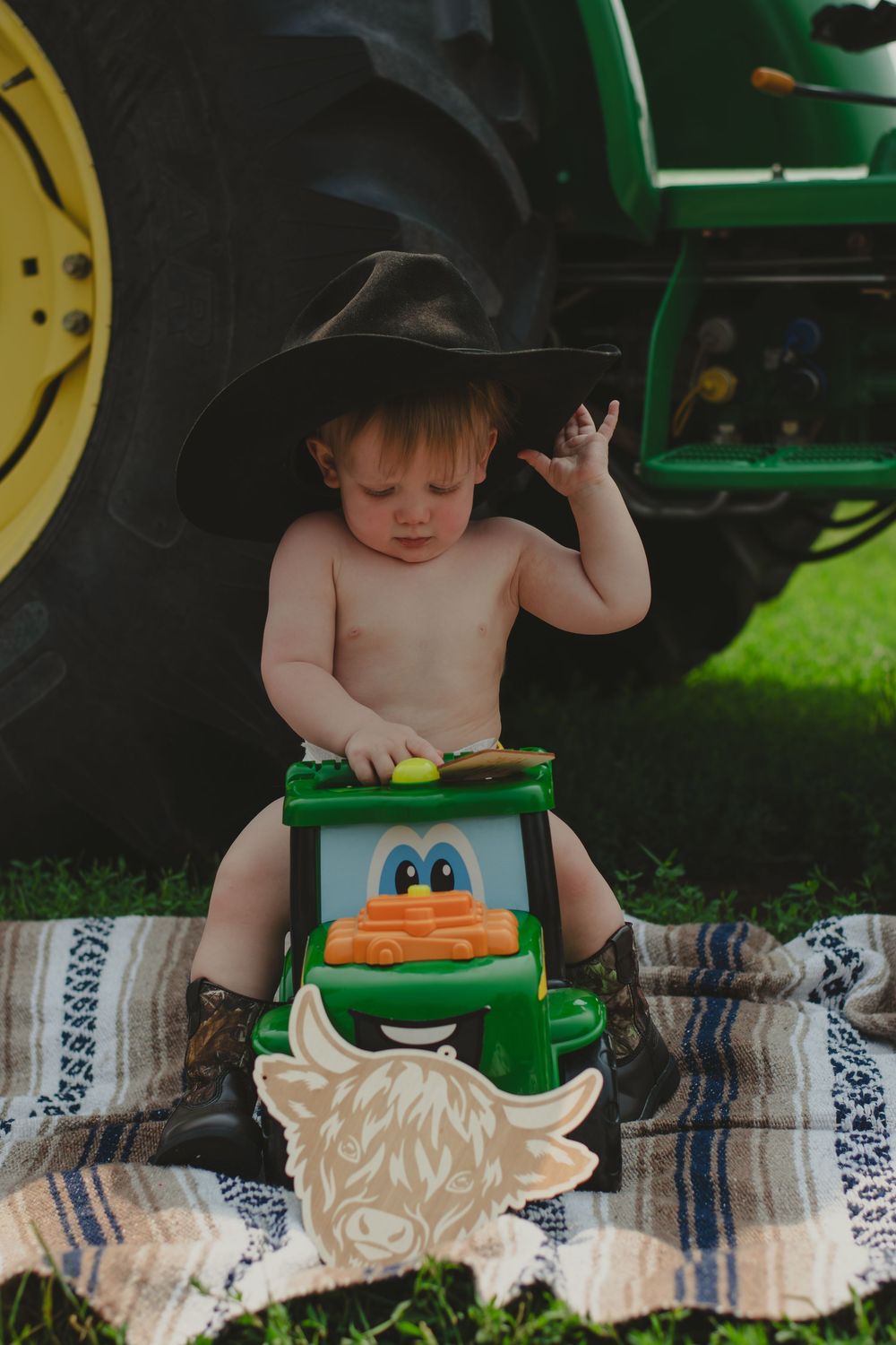 A small child sits on a blanket next to a green tractor on a sunny day.