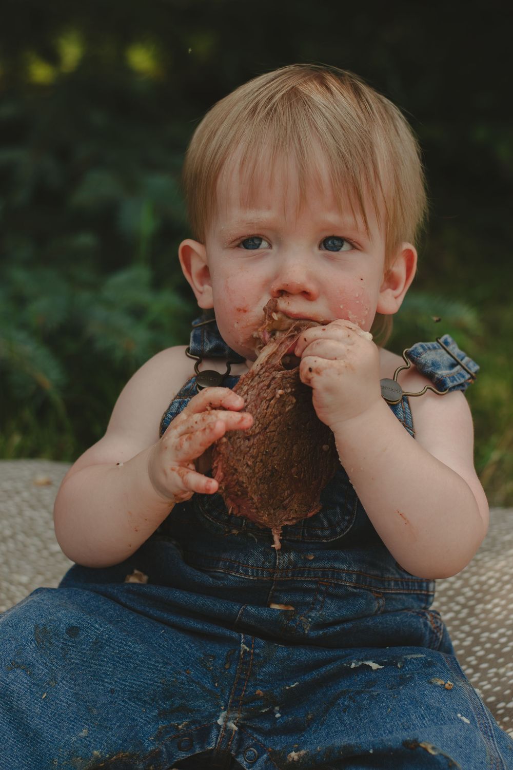 A young child in overalls messily enjoys eating chocolate cake.