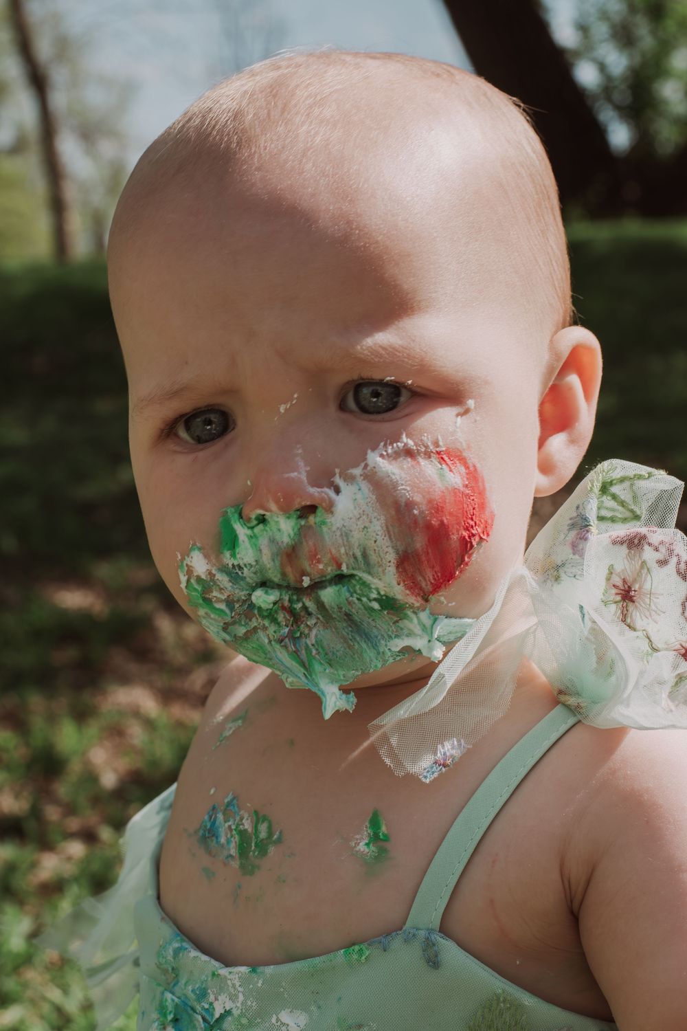 Baby in mint dress has cake frosting smeared playfully across their face during celebration.