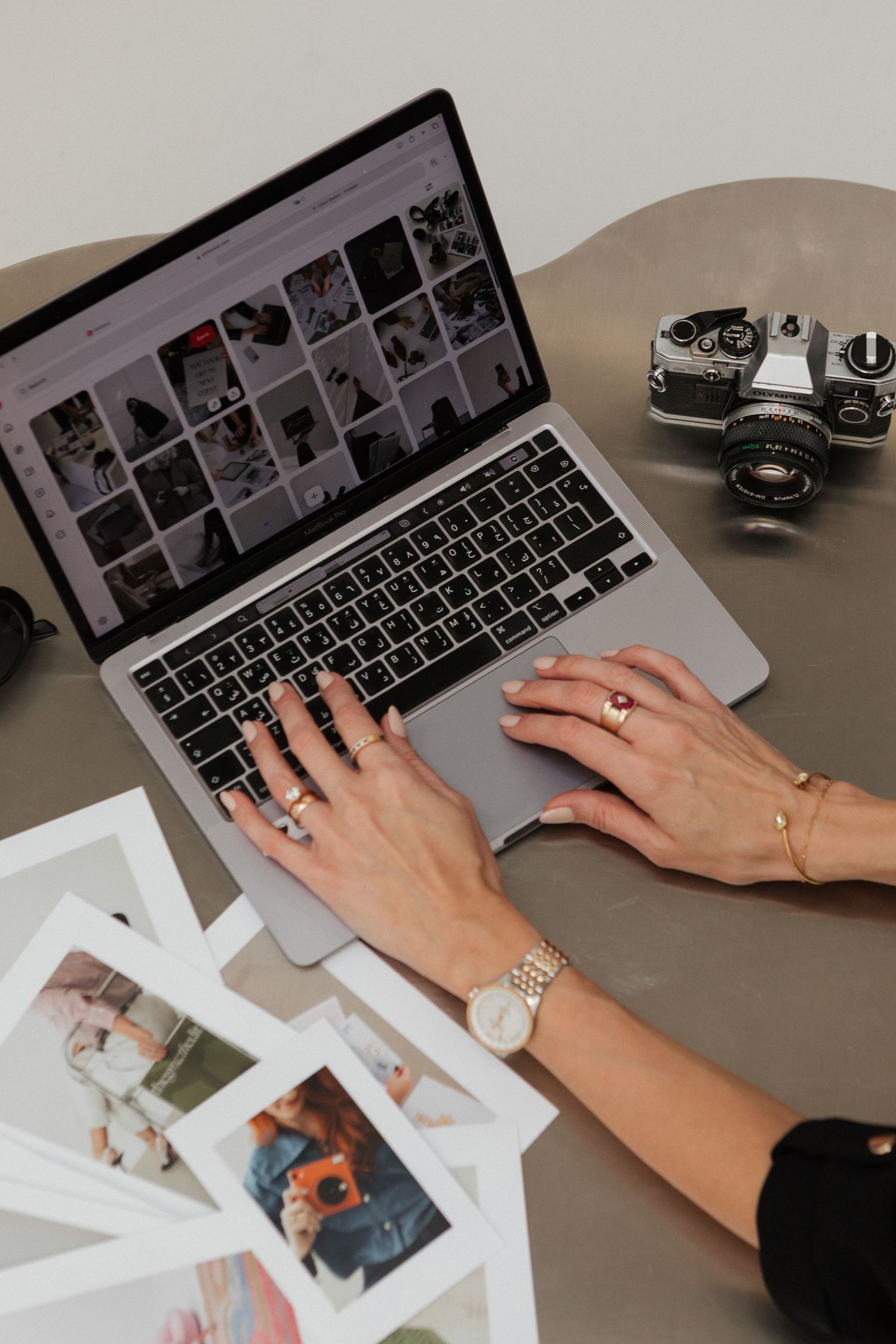 Hands typing on a laptop keyboard while reviewing printed photographs on a desk with a vintage camera nearby.
