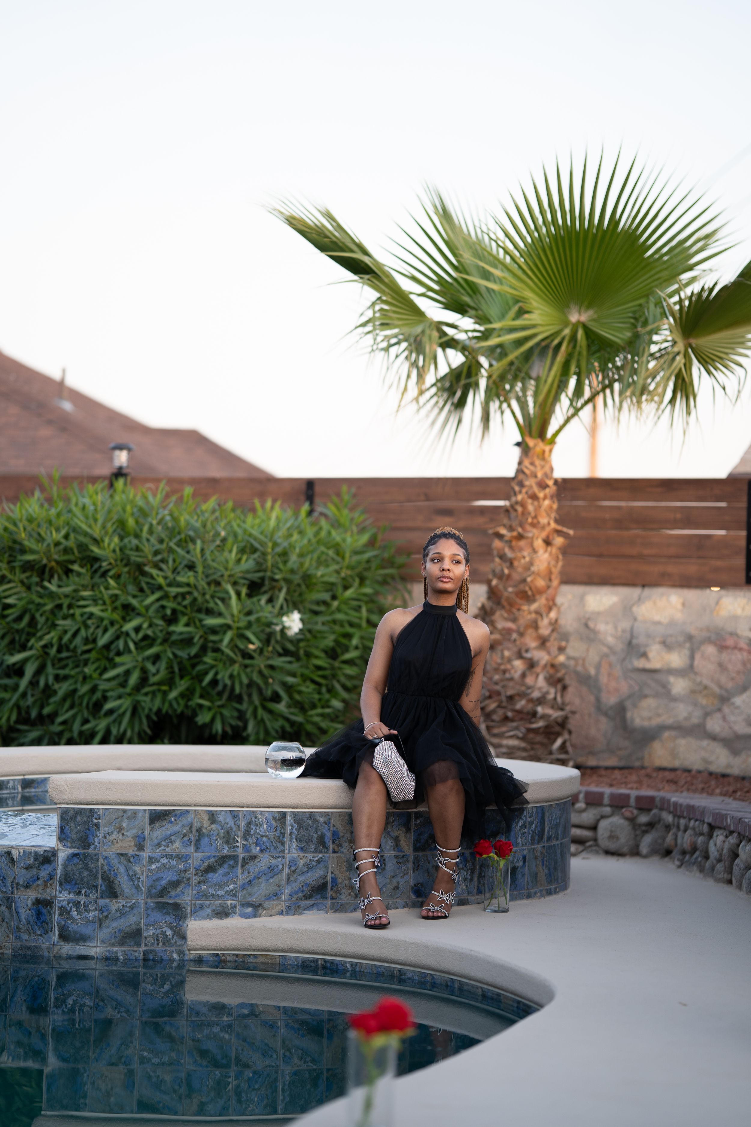 A figure in a black evening dress sits elegantly by a stone-lined pool with palm trees at sunset.