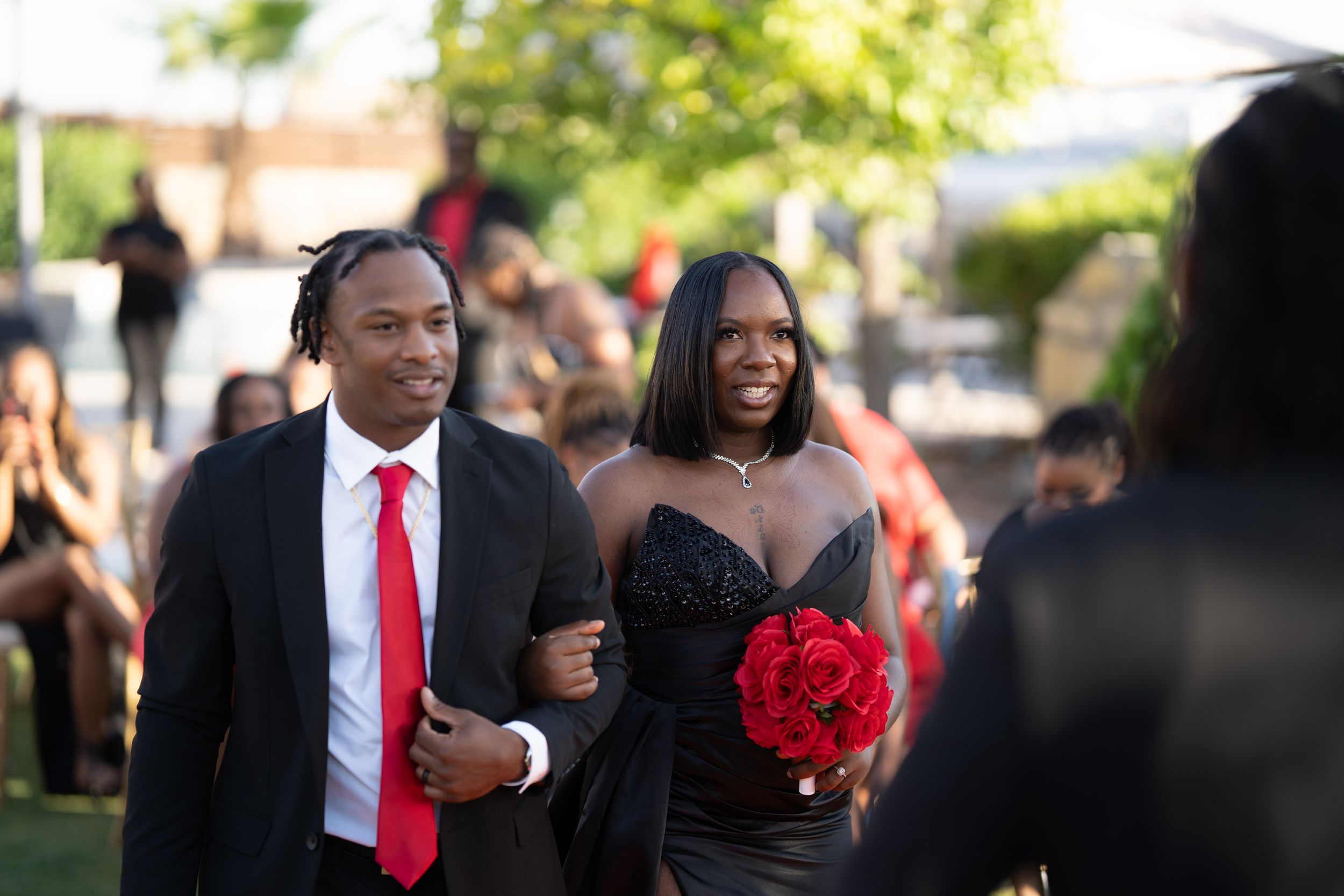 A couple in formal attire smiles together outdoors during a sunny celebration.