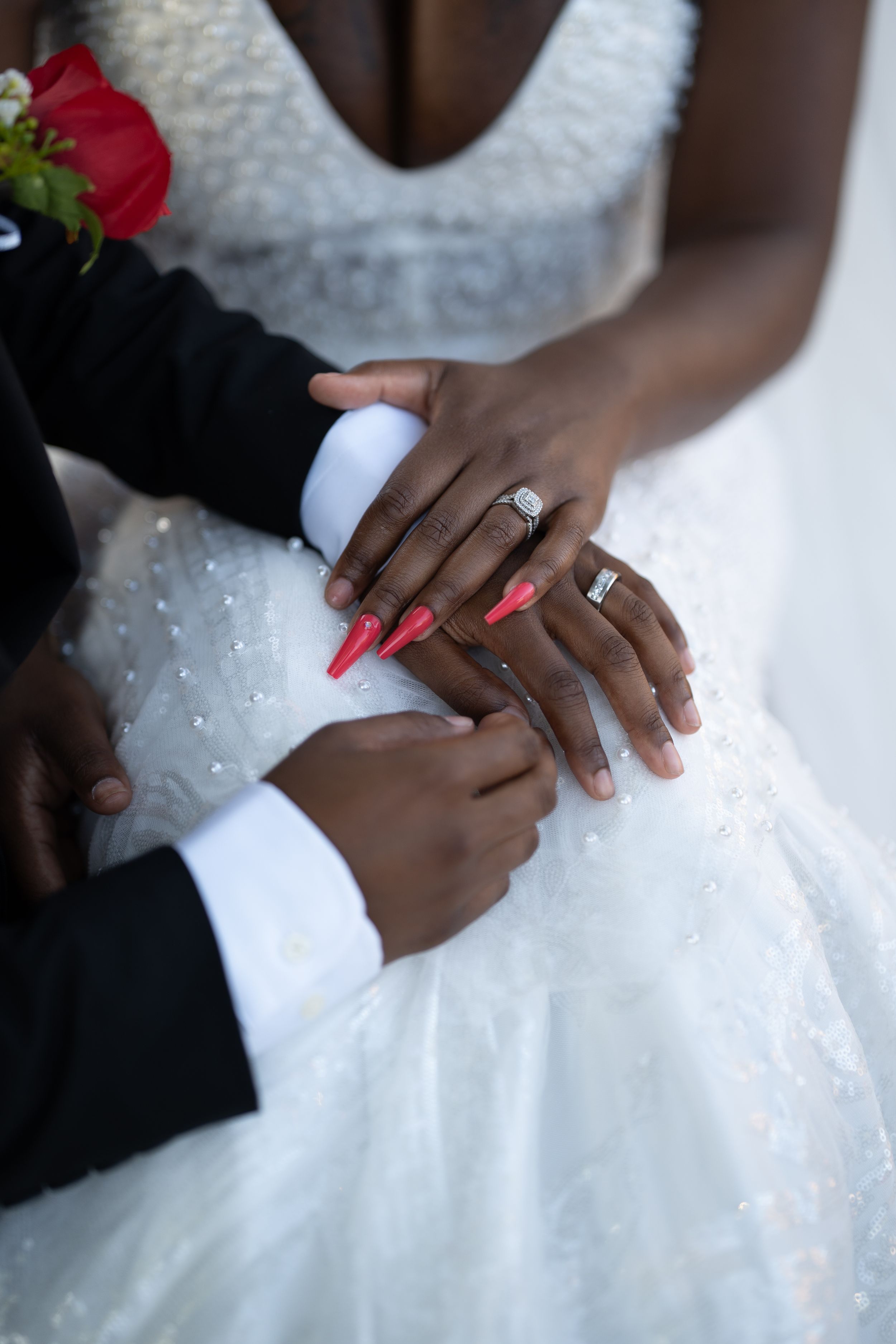 Close-up of hands wearing wedding rings against a white beaded wedding dress with red rose accents.