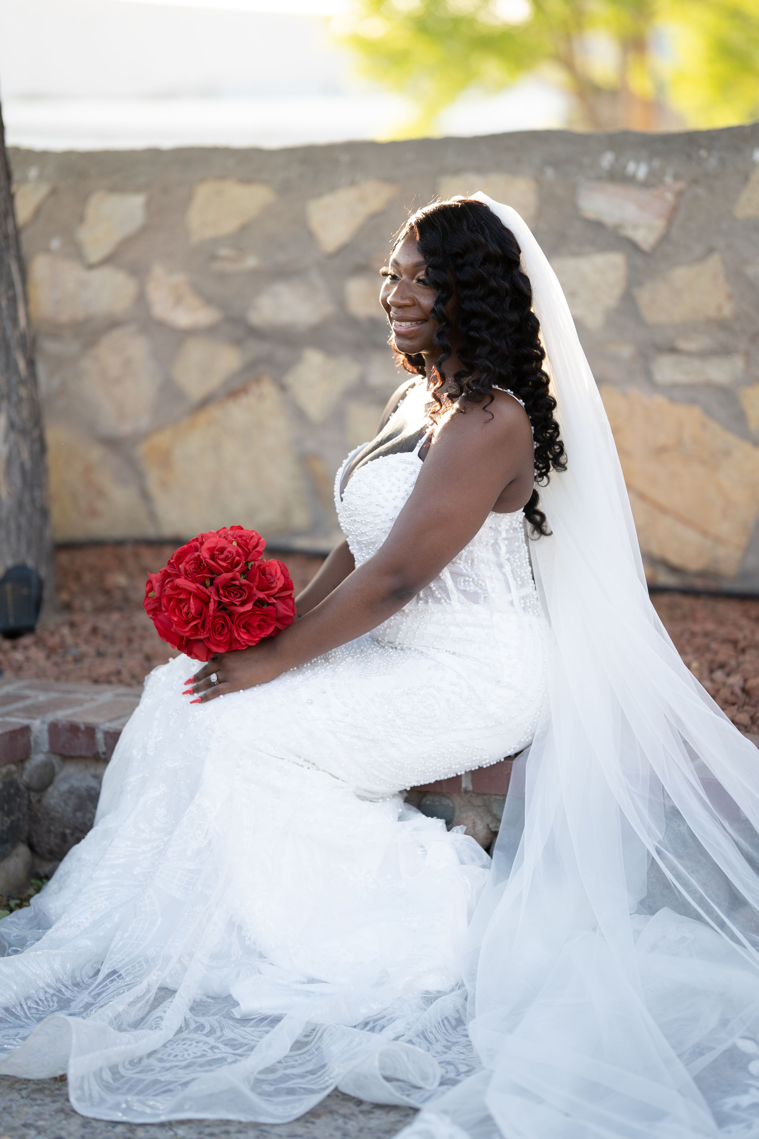 Bride in a flowing white wedding gown sits elegantly with a red rose bouquet against a stone wall backdrop.