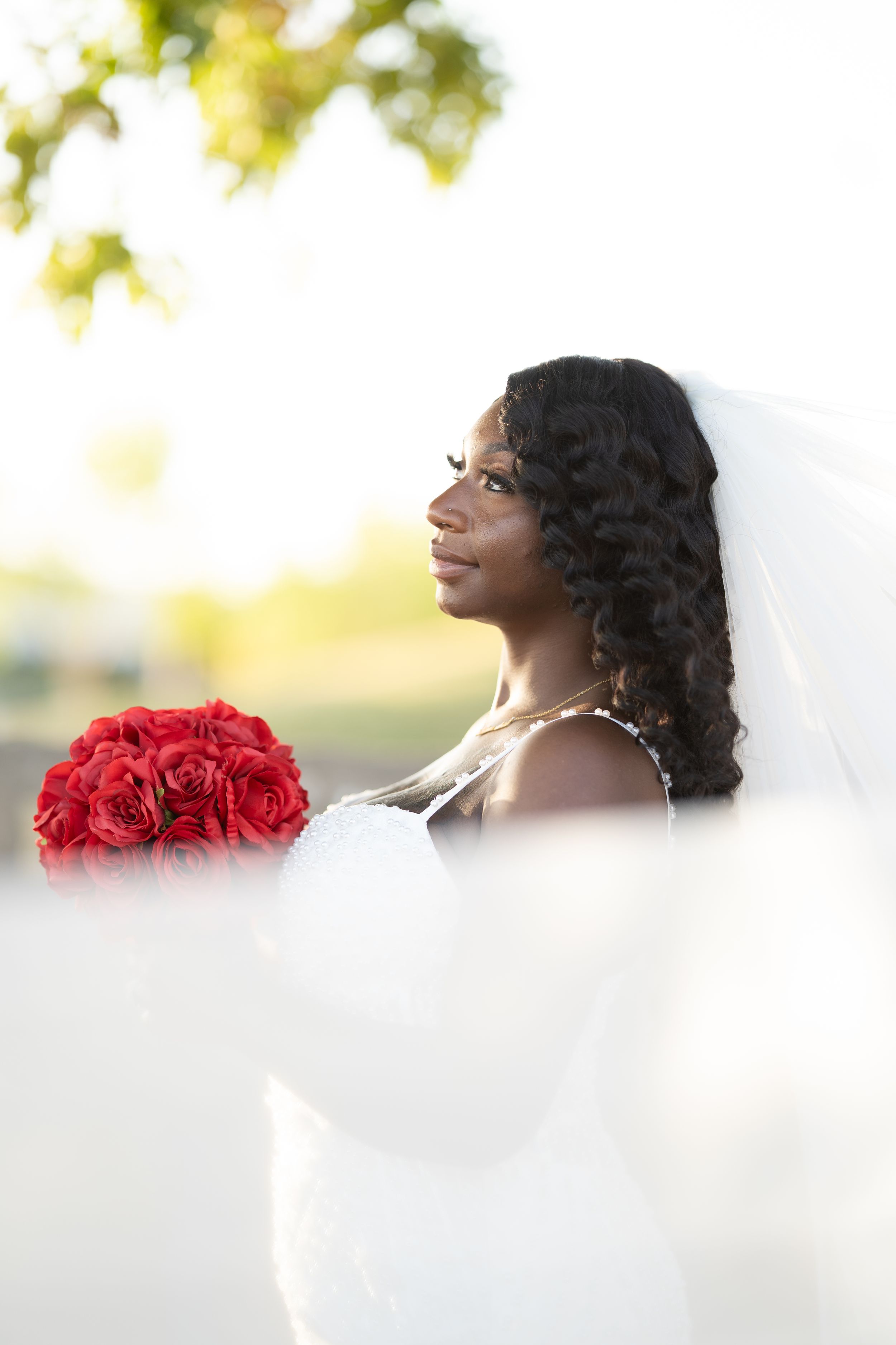 A bride in a white dress poses with a vibrant red rose bouquet against a soft bokeh background.
