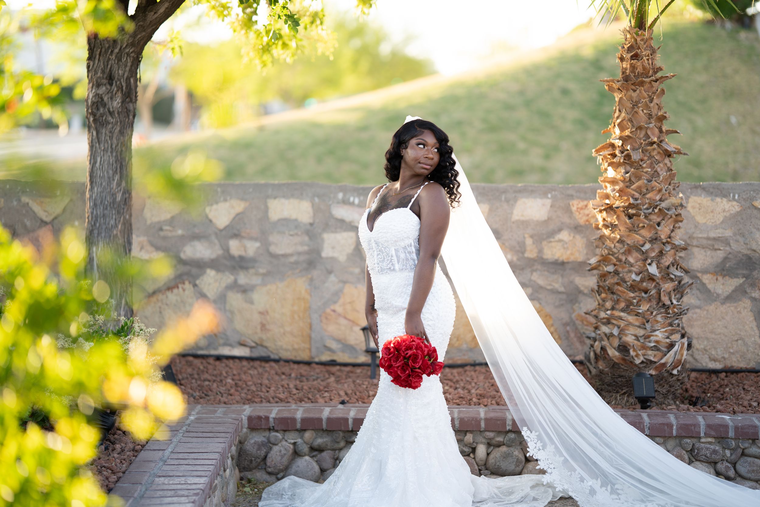 A bride in a white wedding gown with a red sash poses gracefully on stone steps in golden sunlight.