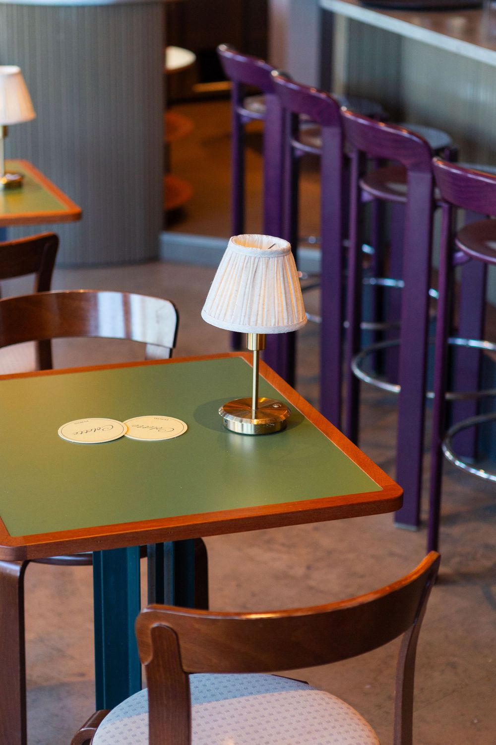 Small restaurant table with green top and miniature table lamp, featuring purple bar stools in background.
