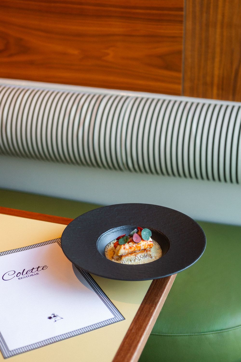Black plate with small appetizer dish and menu on green table surface with striped banquette seating.
