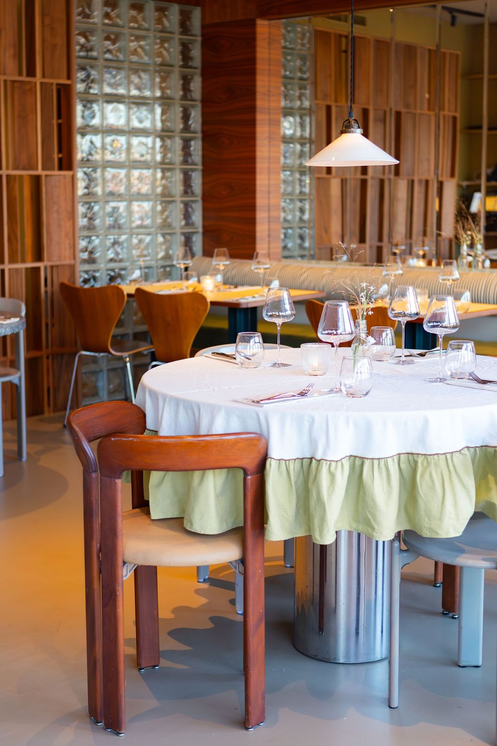 Elegant dining room with round table, white tablecloth, and wooden chairs against a glass block wall.