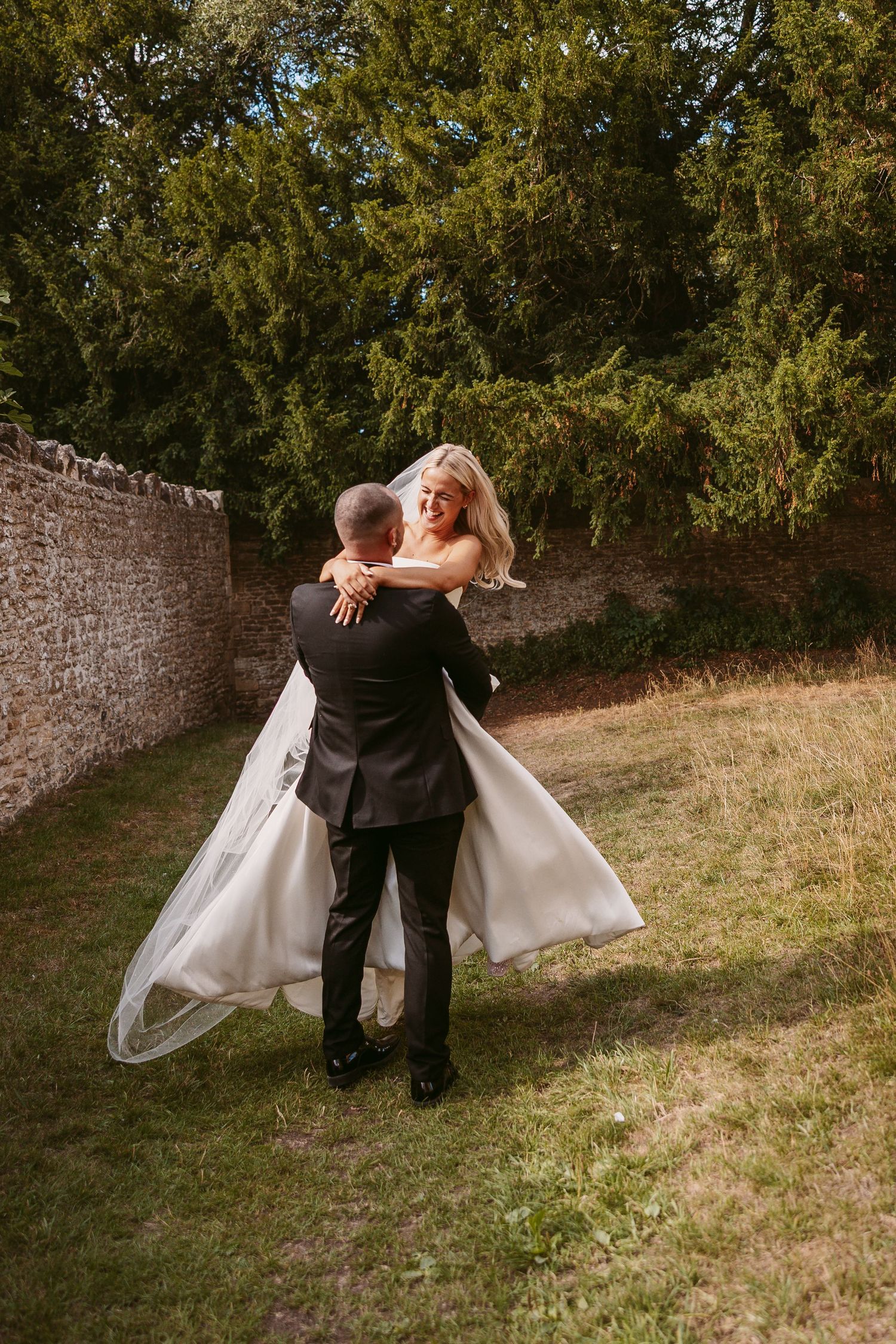 A romantic wedding couple walks along a stone wall on a grassy hillside in matching black and white attire.