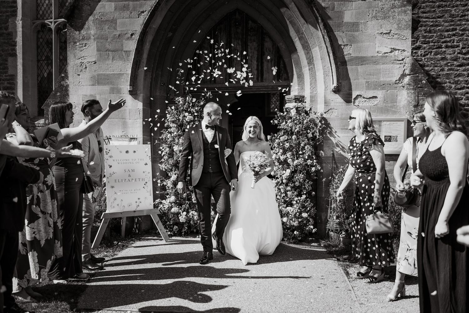 Wedding guests throw confetti as newlyweds exit through a stone archway entrance.