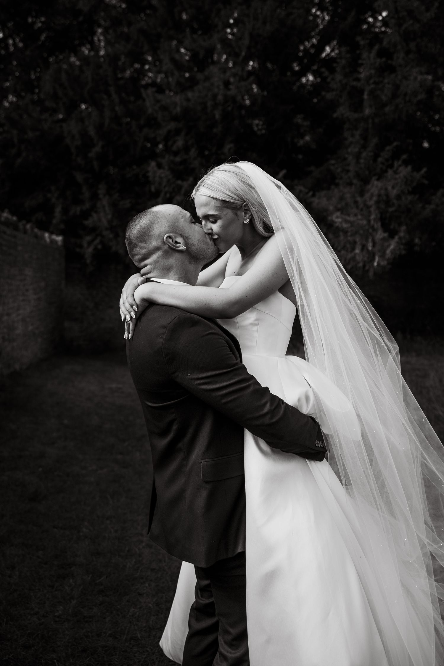 Black and white photo captures an intimate embrace between newlyweds outdoors.