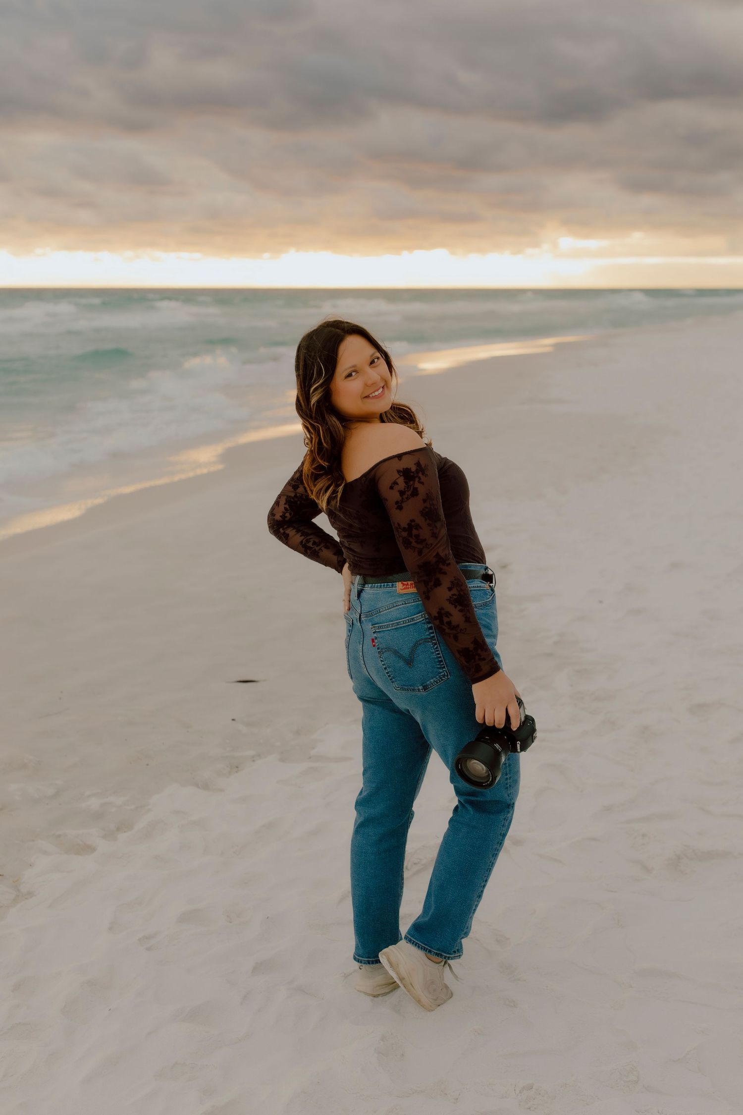 Young woman in dark lace top and jeans holding camera on white sand beach at sunset.