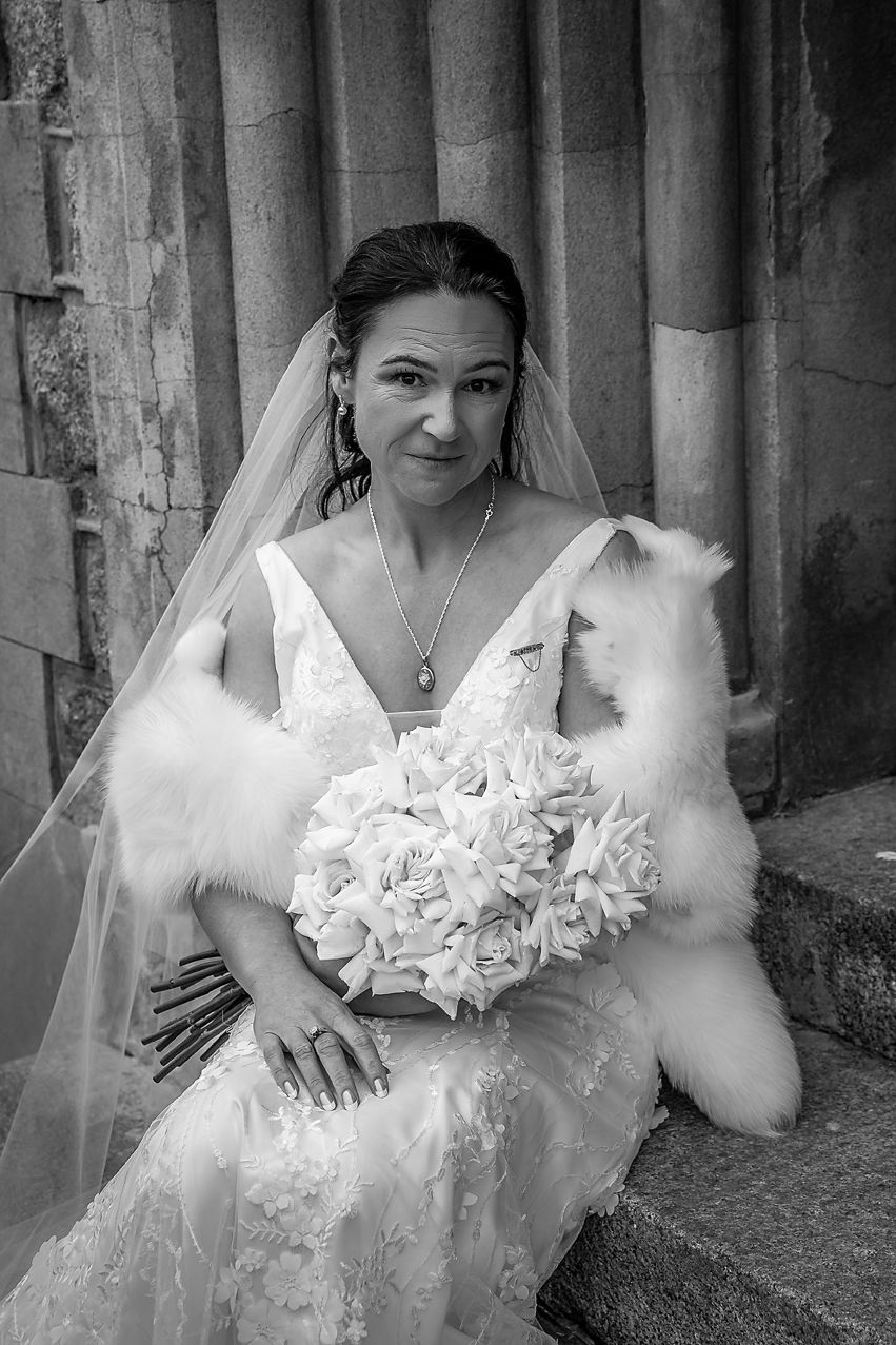 Black and white portrait of a bride in a white wedding dress with fur wrap holding a bouquet of white flowers.