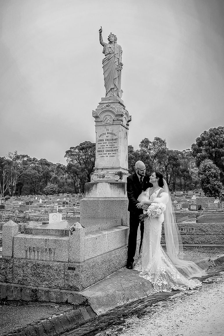 A black and white wedding photo of a couple posing next to a large stone monument in a cemetery.