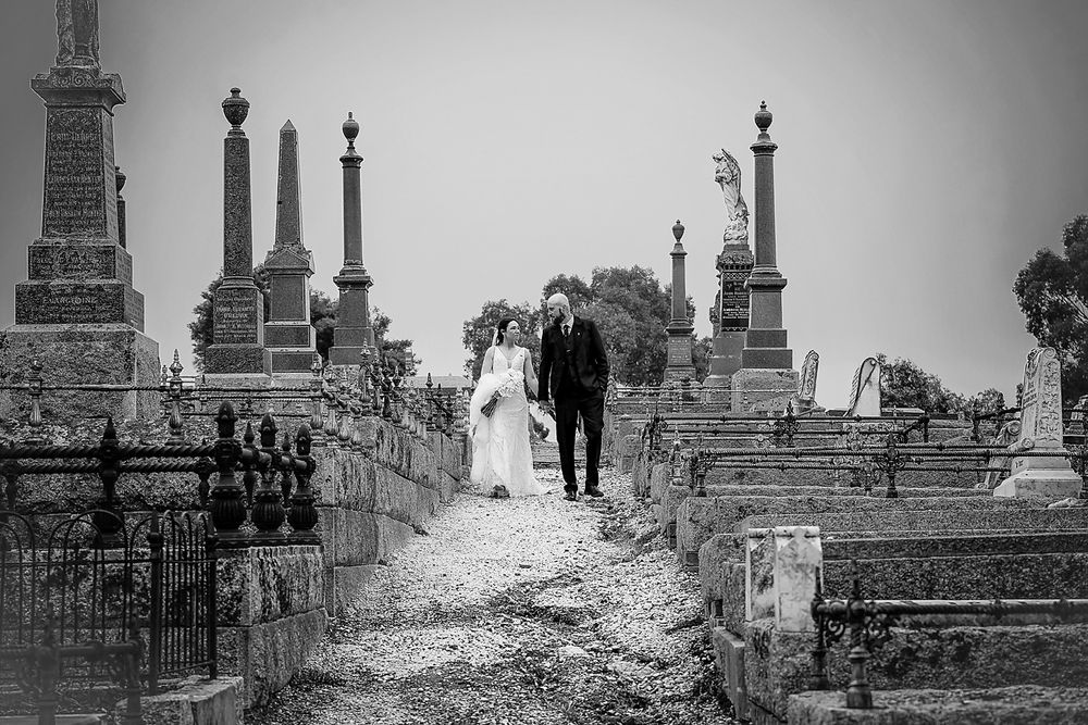 A couple walks between ornate cemetery monuments and gravestones on a misty day in black and white.